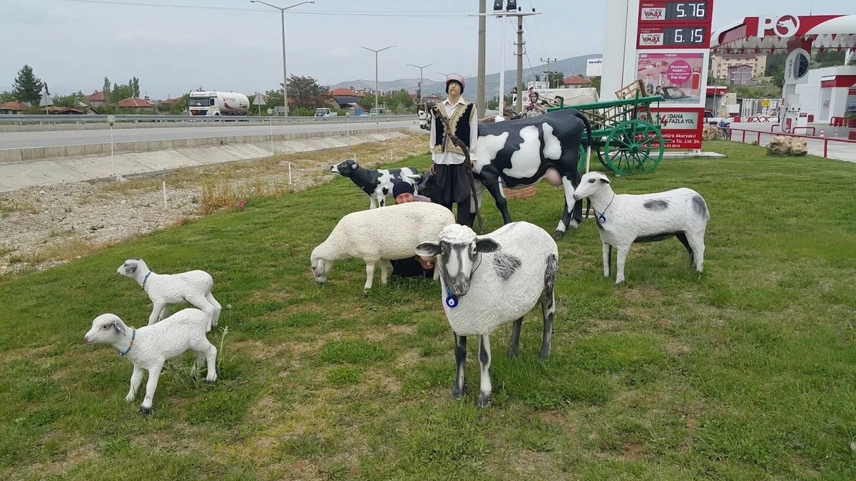 Sığır ve koyun figürlerinin önünde geleneksel kıyafetli bir kişi duruyor, sakin bir doğa manzarası.