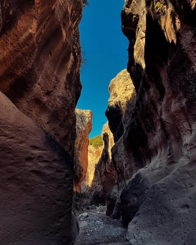 Dar geçit arasında yüksek kayalık duvarlar, pürüzlü yüzeyler ve mavi gökyüzü ile doğal kanyon manzarası.