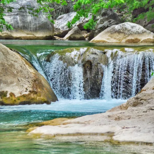 Berrak mavi suyun aktığı şelale, etrafında büyük taşlar ve yeşil ağaçlar var.