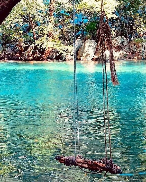 Turkuaz su içinde asılı doğal malzemelerle yapılmış salıncak, yeşil ağaçlar ve büyük kayalarla çevrili.