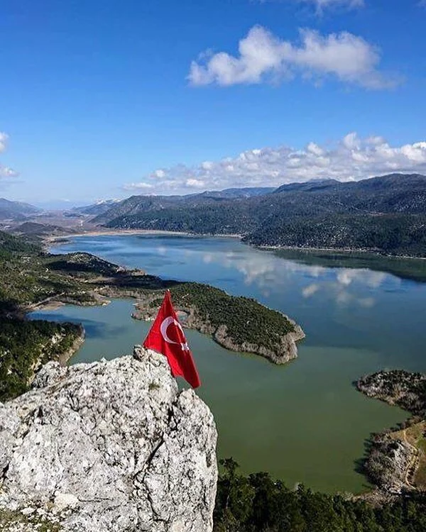 Yüksek kayalığın tepe noktasında dalgalanan Türk bayrağı, arka planda göl ve dağ manzarası.