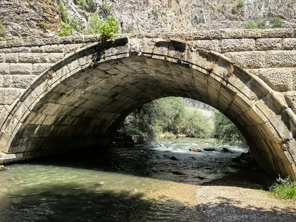 Yüksek taş duvarlarla çevrili doğal manzarada büyük kemerli köprü ve berrak nehir görünmektedir.