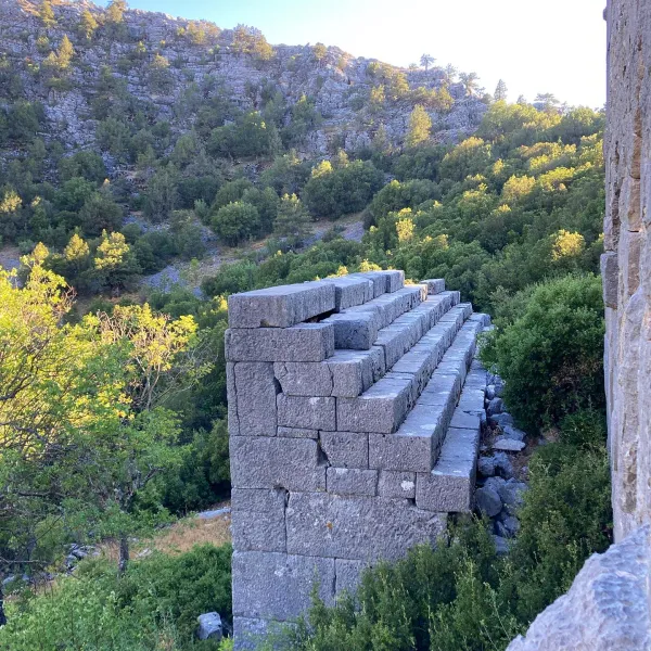 Taşlardan yapılmış harabe bir yapının yan duvarı, arka planda yeşil dağ manzarasıyla birlikte görünüyor.
