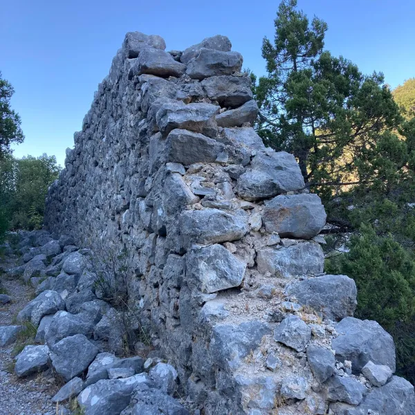 Doğal ortamda yer alan taş duvar, arka planda yeşil ağaçlar ve mavi gökyüzü ile tarihi bir yapıyı yansıtıyor.
