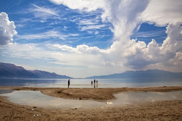 Eğirdir Gölü'nde plajda insanlar suya ilerliyor, dağlar ve bulutlar arka planda.