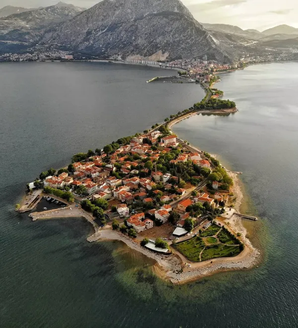 Su ile çevrili yarımada, yeşil alanlar ve kırmızı çatılı evler, arka planda dağlar ve mavi gökyüzü