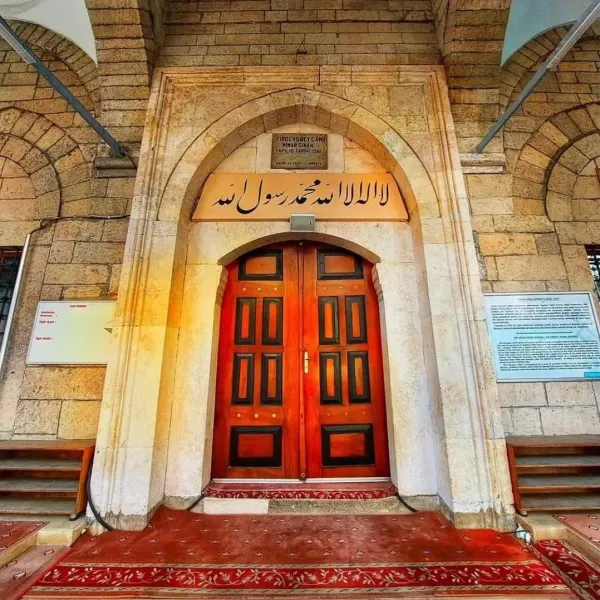 Ahşap siyah kapı, altın menteşe ve Arapça yazılarla tarihi bir caminin girişini gösteriyor.