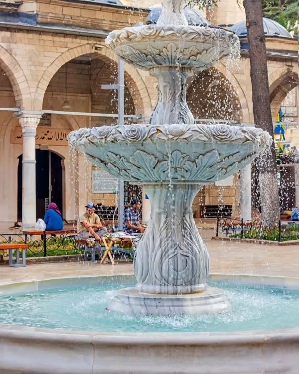 Mermer çeşme, yeşil bitkilerle dolu avlu ve taş yapılarla çevrili.