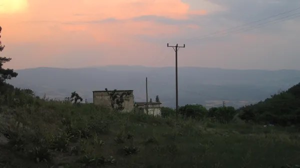 Kuyucak Köyü'nde ot ve çalılarla kaplı zemin, eski yapı ve dağ silsilesi ile pastel tonlarda gün batımı.