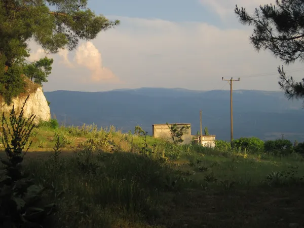 Yeşil bitki örtüsü arasında iki bina ve dağlar, pembe tonlarda bulutlu gökyüzü.