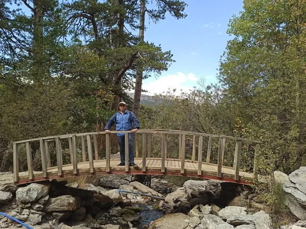 Ahşap köprüde duran mavi giysili kişi, yeşil ağaçlar ve su kaynağıyla çevrili huzurlu doğal manzara.