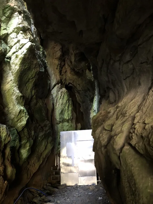 Büyük bir doğal mağara, pürüzlü duvarlar ve metal kapı ile ahşap merdivenler içeren gizemli bir atmosfer.