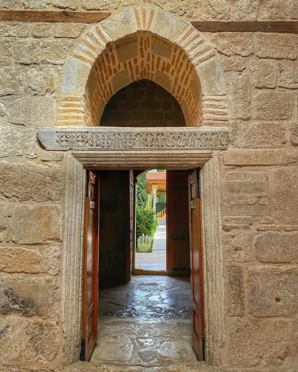 Taş duvarlarla çevrili, süslü kapı açılmış, yeşilliklerle dolu bir alan görünüyor.