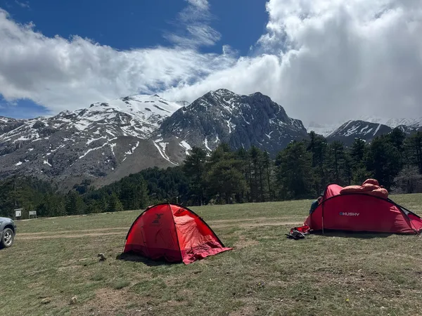 Yüksek dağlarla çevrili yeşil alanda iki kırmızı çadır ve ağaçlar.