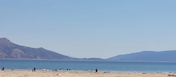 Geniş plajda insanlar yürüyüş yapıyor ve denizde yüzüyor, arka planda dağlar uzanıyor.