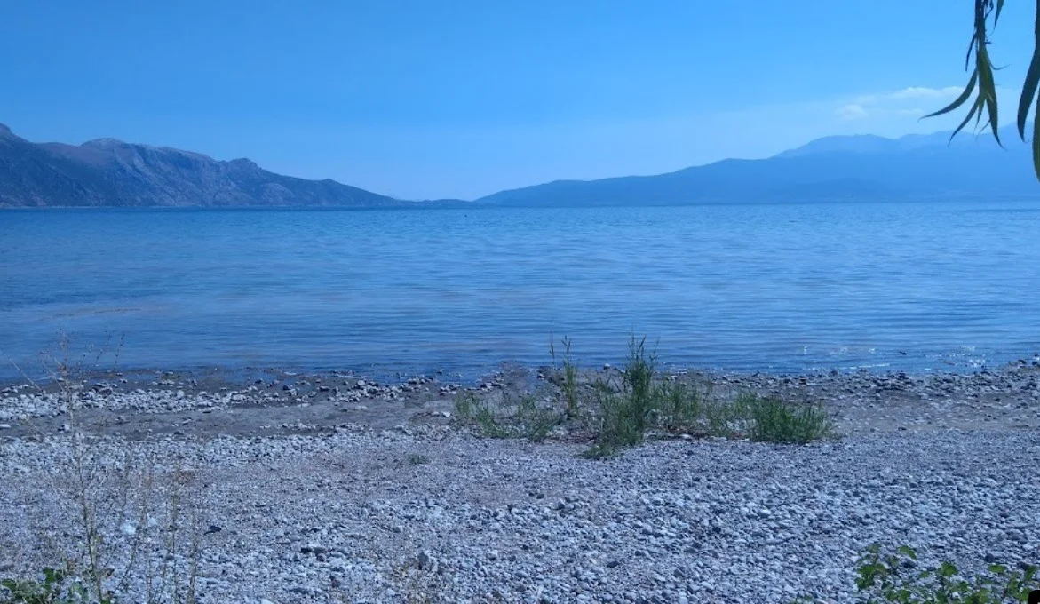 Sakin göl manzarasında hafif taşlı bir plaj ve dağ silueti
