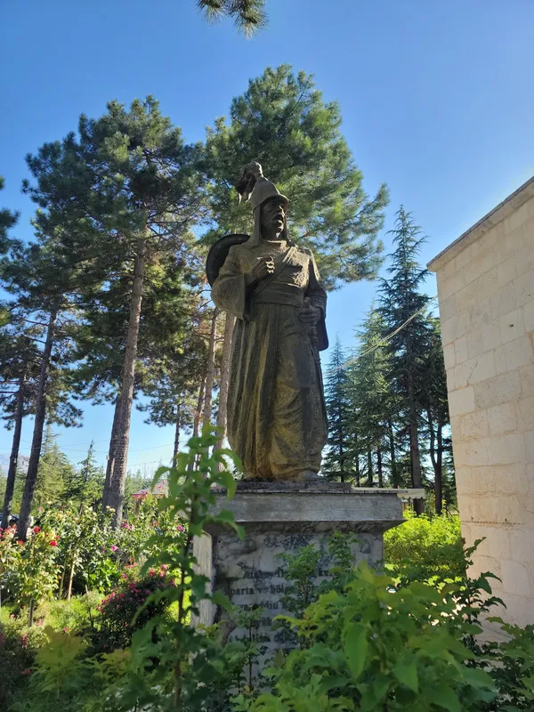 Geleneksel kıyafetli bir heykel, yeşil ağaçlarla çevrili alanında duruyor.