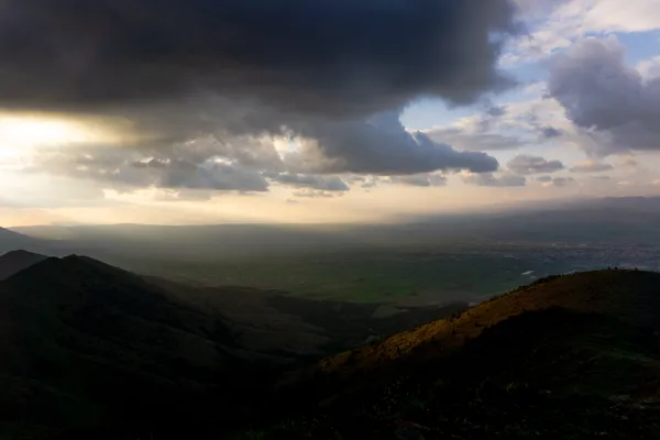 Dağlık manzarada bulutlu hava ve yeşil tarım arazileri görünmekte.