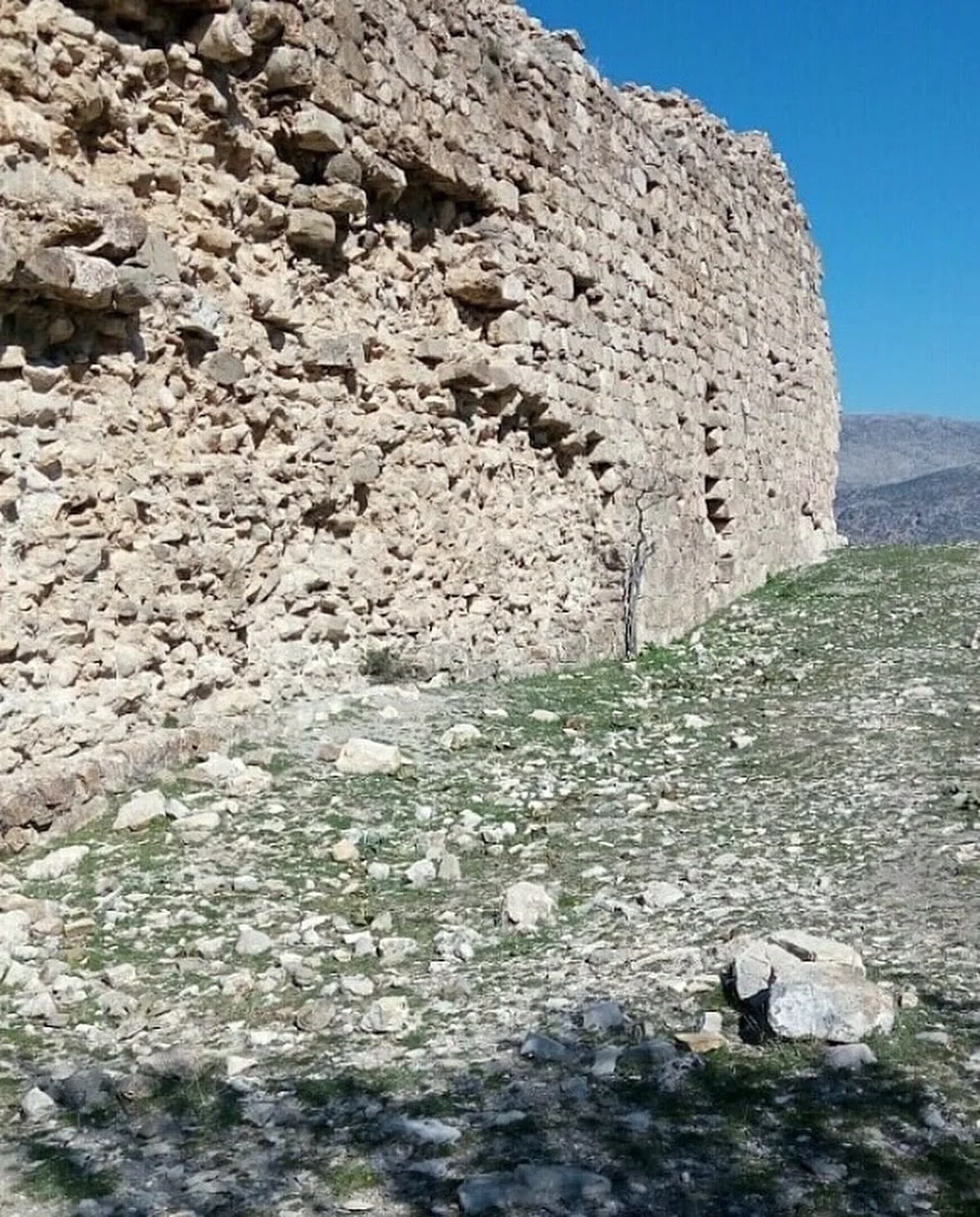 Taş duvar, mavi gökyüzü ve dağlarla çevrili doğal alan.