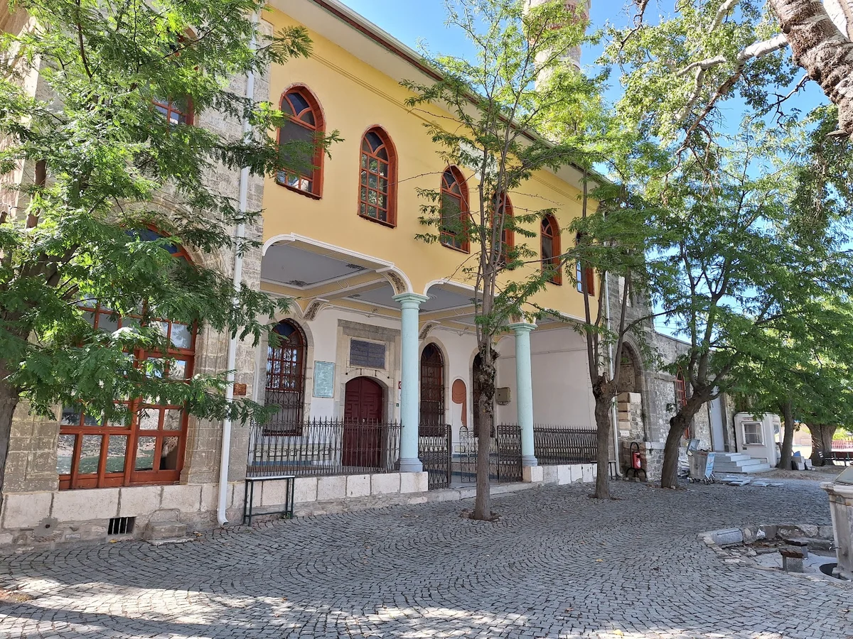 Taş yapının ön planda olduğu sokak manzarası, sarı pencereler ve yeşil ağaçlar ile çevrili.
