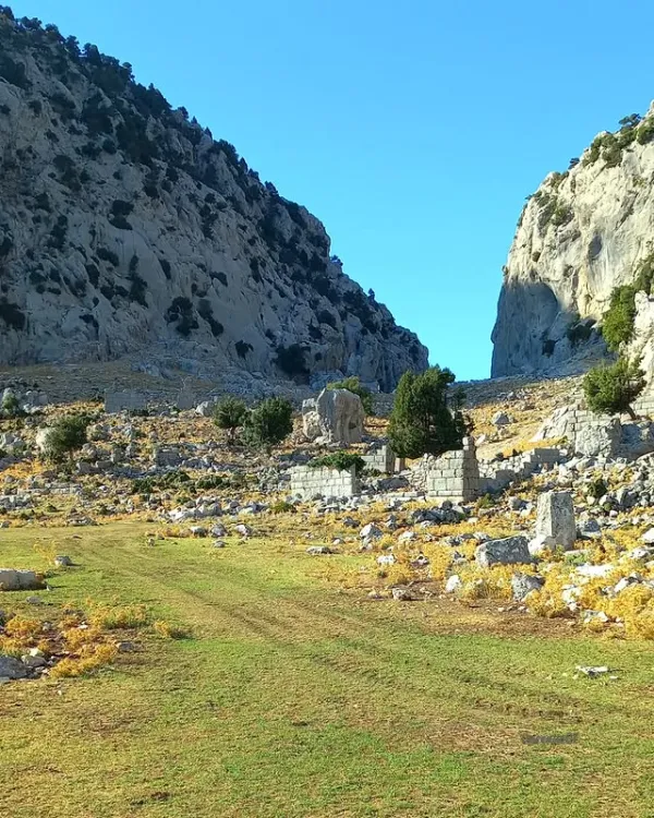 Yüksek dağlarla çevrili açık alan, sarı ve yeşil bitki örtüsü, taş kalıntıları ve çalılar ile doğal bir manz