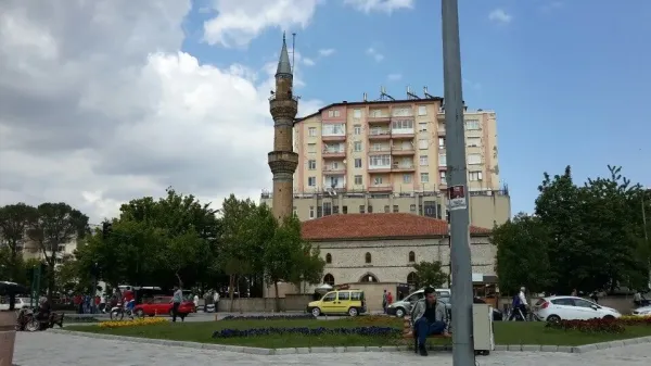 Tarihi cami minaresi, yeşil ağaçlar ve modern apartmanlarla çevrili hareketli bir meydan.