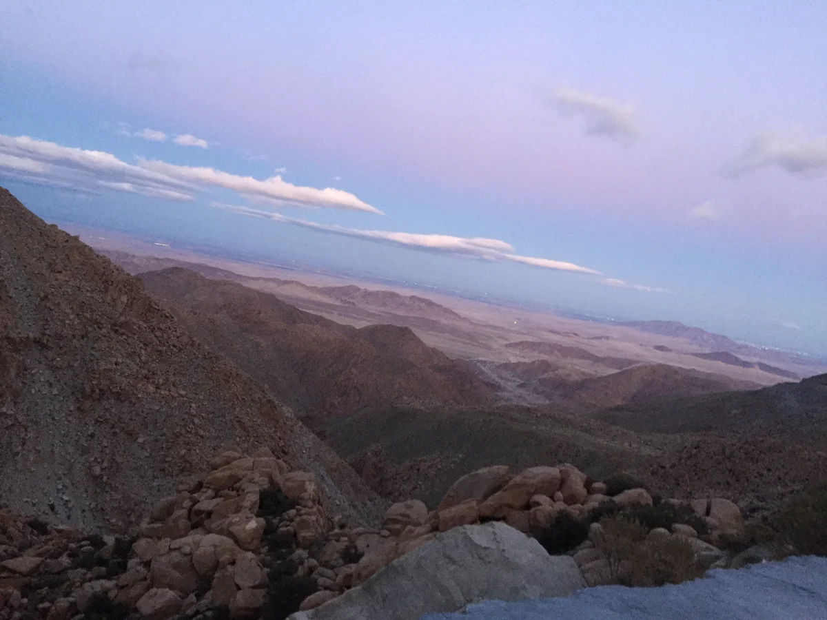 Geniş dağlık alan ve vadilerin tepe görünümü, pembe ve mavi bulutlarla dolu gökyüzü.