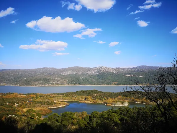 Geniş göl manzarası, yeşil dağlar ve açık mavi gökyüzü ile huzur verici bir doğa görüntüsü.