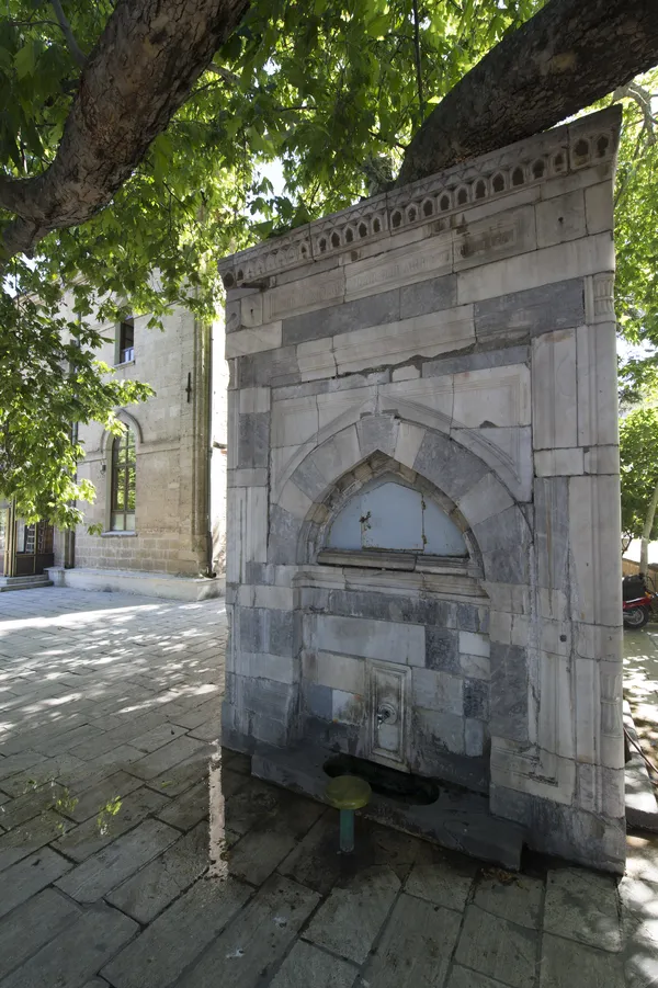 Taş bir çeşme, oymalı detaylar ve tarihi yapılarla çevrili.