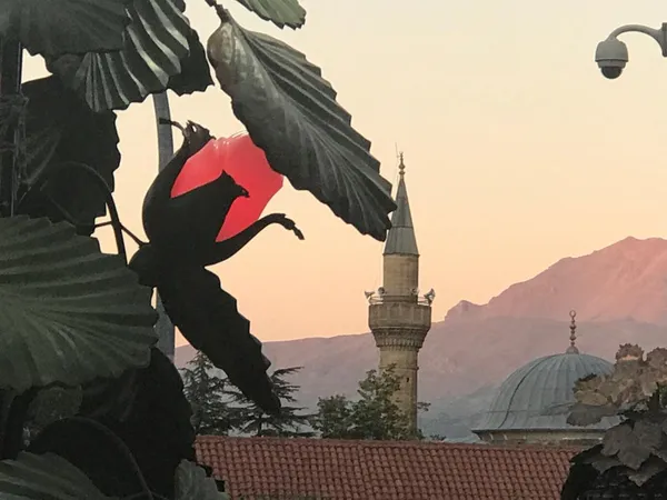 Kutlu Bey Camii minaresi ve kubbesi, yeşil yapraklar ve kırmızı çiçekle çevrili huzur dolu bir manzara.
