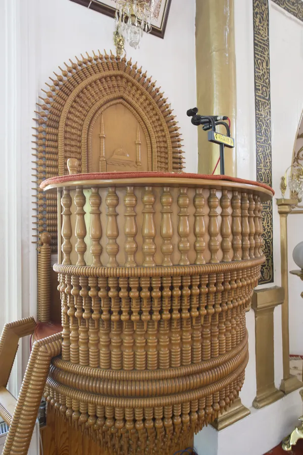 Detaylı işlemeli ahşap minber, yuvarlak sütunlar ve aydınlatma unsurlarıyla süslü.