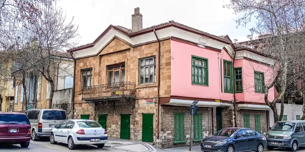 Kahverengi taşlar ve pembe cephe ile tarihi bir bina, araçlar ve ağaçlarla çevrili.