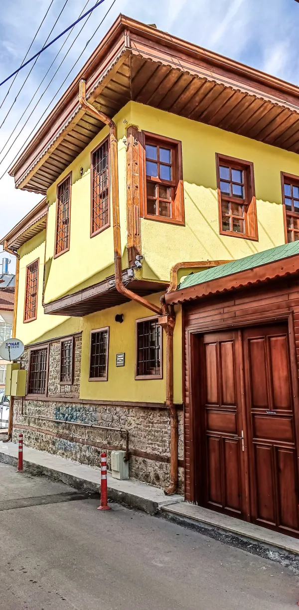 Sarı dış cepheli, ahşap pencereli tarihi bina, park alanı ve otobüs duraklarıyla çevrili.