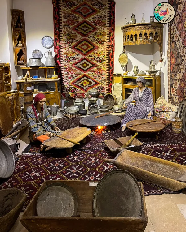 Geleneksel yaşam alanında, elbiseleriyle iki kadın ve ahşap masalar yer alıyor.