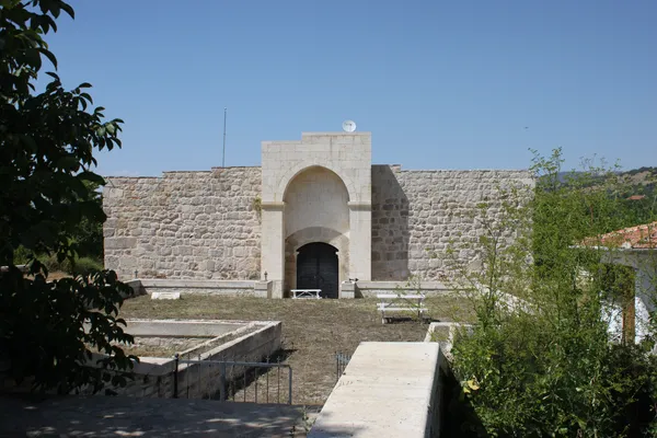 Kalın taşlardan yapılmış surun önünde büyük bir kapı ve çevresinde beyaz masa ve sandalyeler.