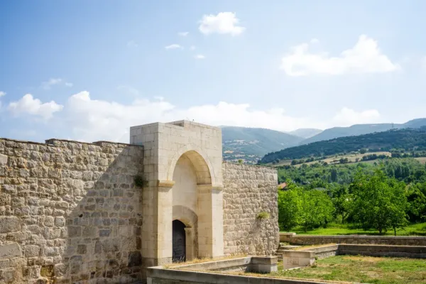 Ertokuş Hanı'nın önünde büyük kemer, arka planda yeşil dağlar ve mavi gökyüzü görünmekte.