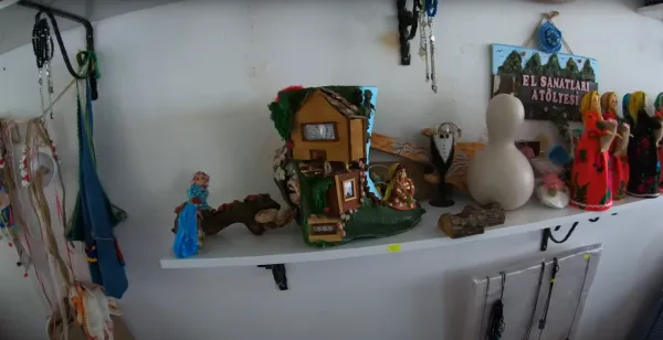Raf üzerinde çeşitli el yapımı sanat eserleri ve renkli çizmeye benzer ev figürü sergileniyor.