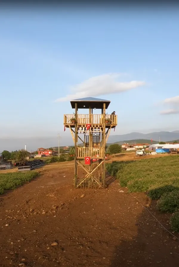 Güneykent kasabasında ahşap gözetleme kulesi, dağ manzarası ve yeşil bitkilerle çevrili.