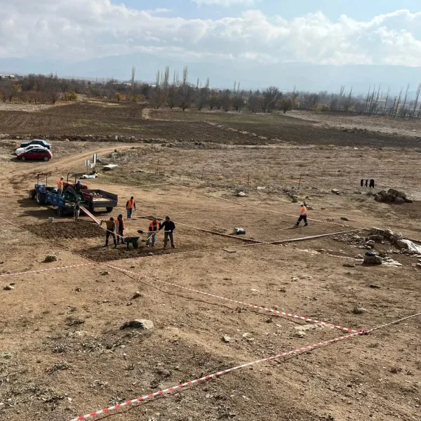 Kazı alanında güvenlik yelekleri giymiş insanlar çalışıyor, arka planda araçlar ve doğal manzara var.