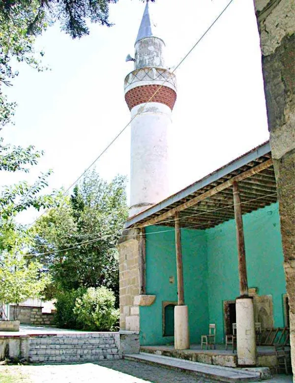 Mavi-yeşil duvara sahip cami ve kırmızı-beyaz desenli minaresi, çevresindeki yeşil ağaçlar ile birlikte