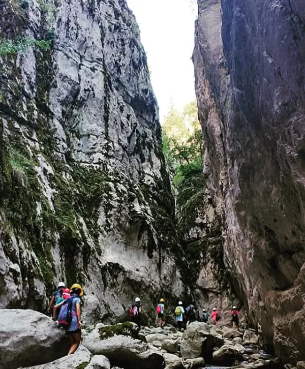 Yüksek kayalıklar arasında yürüyüşçüler ilerliyor, gökyüzü açık mavi.