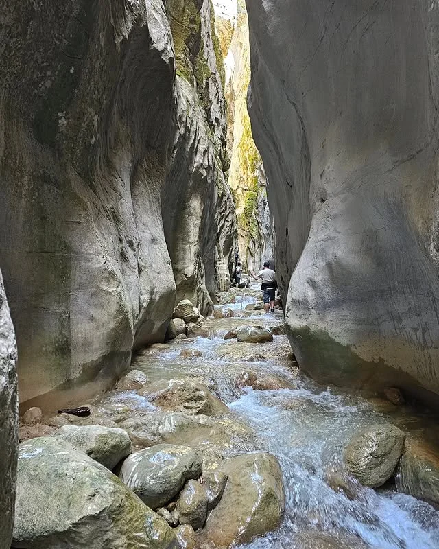 Dar bir kanyonun içinde akışkan bir dere ve yürüyen insanlar görünmekte.