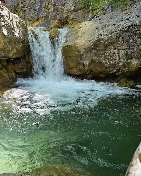 Küçük bir şelalenin kayalar arasından düştüğü, yeşilliklerle çevrili doğal bir manzara.
