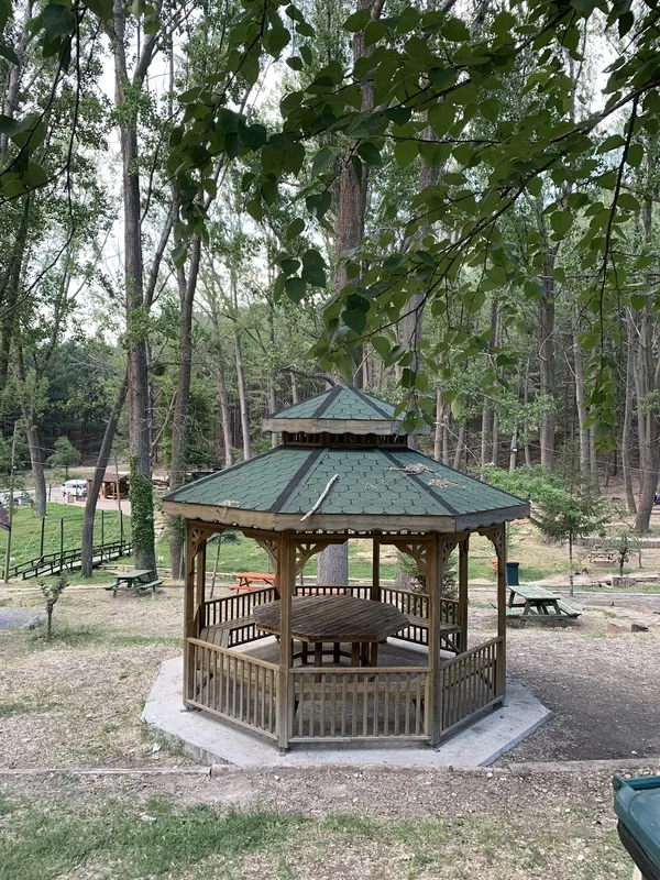 Ağaçlarla çevrili ahşap gazebo, yeşil çatı ve piknik masalarıyla doğal bir ortam sunuyor.