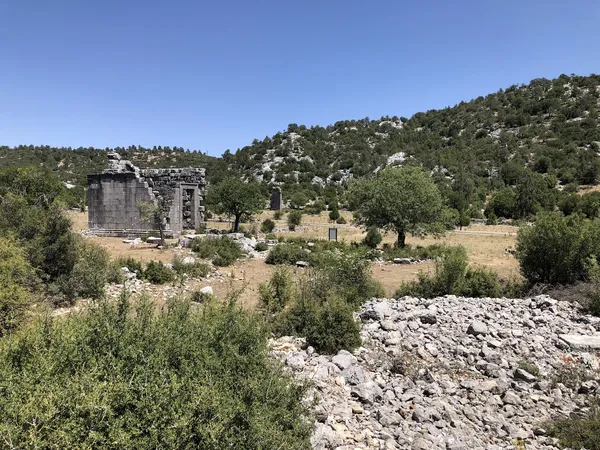 Yeşil arazide taş kalıntıları ve antik yapılar, arka planda dağ silueti ve mavi gökyüzü ile birleşiyor.