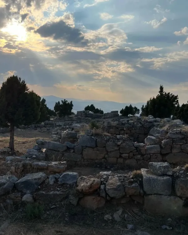 Taşlardan yapılmış tarihi kalıntılar, çam ağaçları ve dramatik gökyüzüyle manzara.