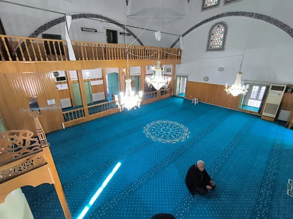 Büyük bir cami iç mekanında mavi halı, yüksek tavan ve ibadet eden bir kişi görünmekte.