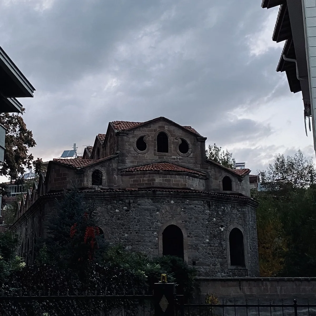 Tarihi taş yapı, yuvarlak pencereleri ve yeşil bitkilerle çevrili.