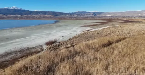 Açık mavi suya sahip göl, çevresinde dağlar ve ağaçlık alanlar ile huzurlu bir manzara sunuyor.