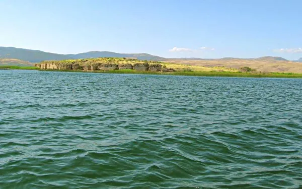 Sakin su yüzeyi, hafif dalgalar ve yeşil tepelerle mavi gökyüzü.
